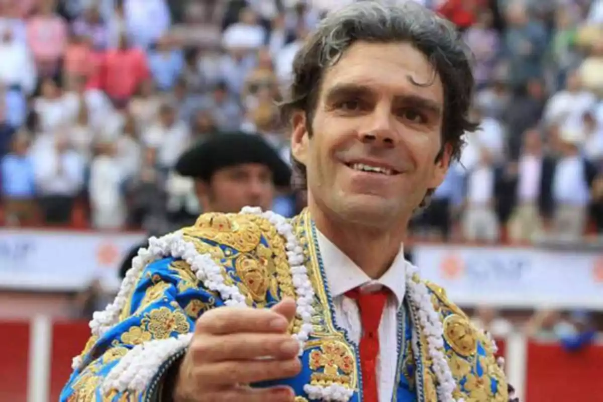 Un torero sonriente con traje de luces azul y dorado en una plaza de toros llena de espectadores.