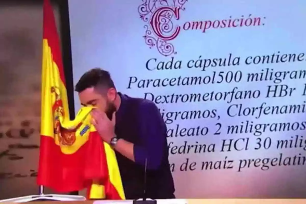Un hombre con barba besa una bandera de España mientras está de pie frente a una pantalla que muestra la composición de un medicamento.