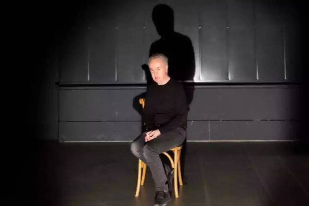 Persona sentada en una silla de madera en una habitación oscura con una sombra proyectada en la pared detrás de él.