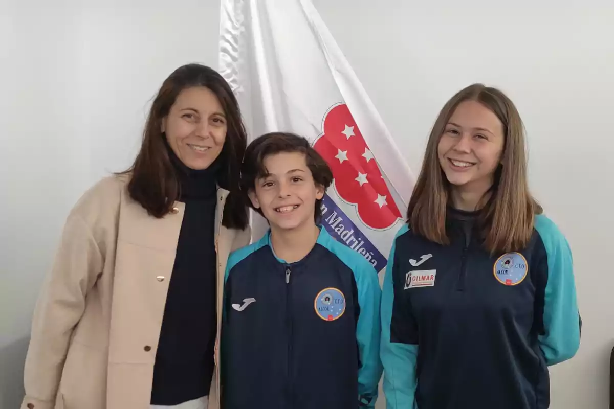 Tres personas posando frente a una bandera con un escudo.