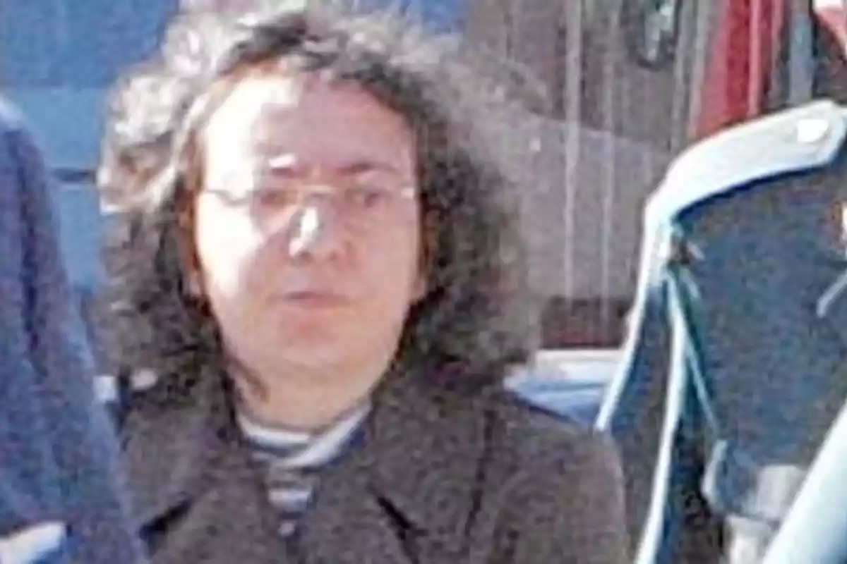 Una persona con cabello rizado y gafas, vestida con un abrigo oscuro, está de pie al aire libre con un fondo borroso que incluye una chaqueta azul colgada.