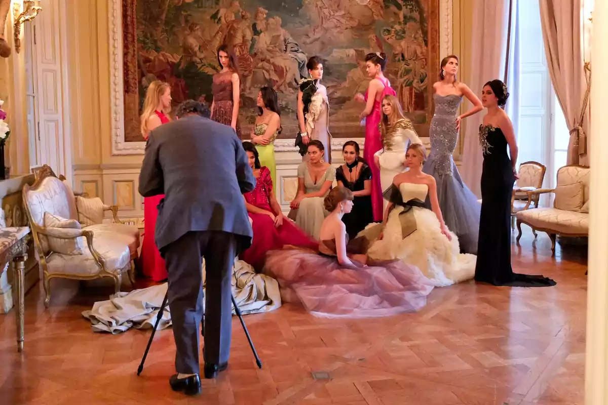 Un fotógrafo captura a un grupo de modelos elegantemente vestidas en un salón decorado con un gran tapiz en la pared.