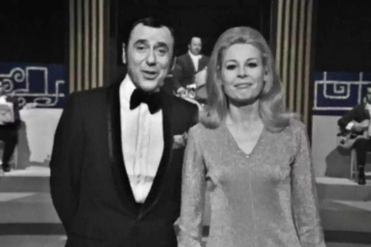 Una pareja de presentadores en un programa de televisión en blanco y negro con músicos de fondo.