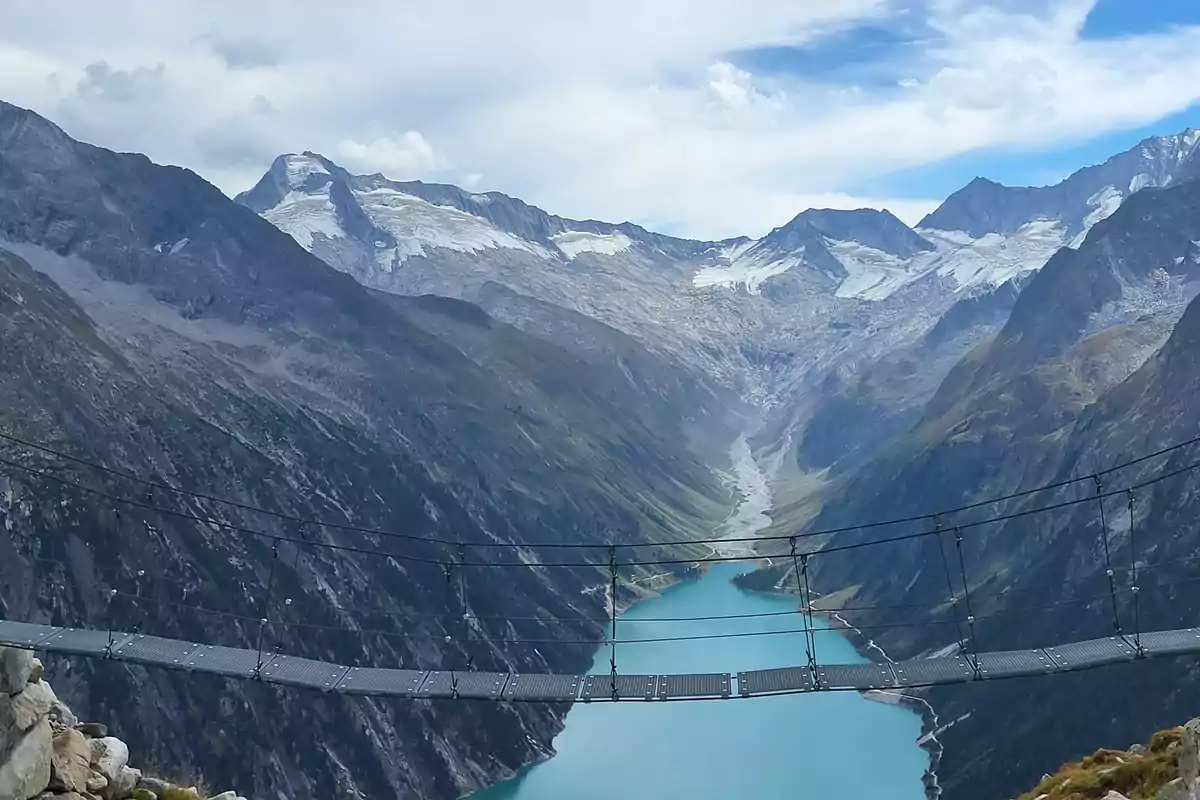 Puente colgante sobre un valle montañoso con un lago de agua turquesa y montañas nevadas al fondo Puente colgante sobre un valle montañoso con un lago de agua turquesa y montañas nevadas al fondo