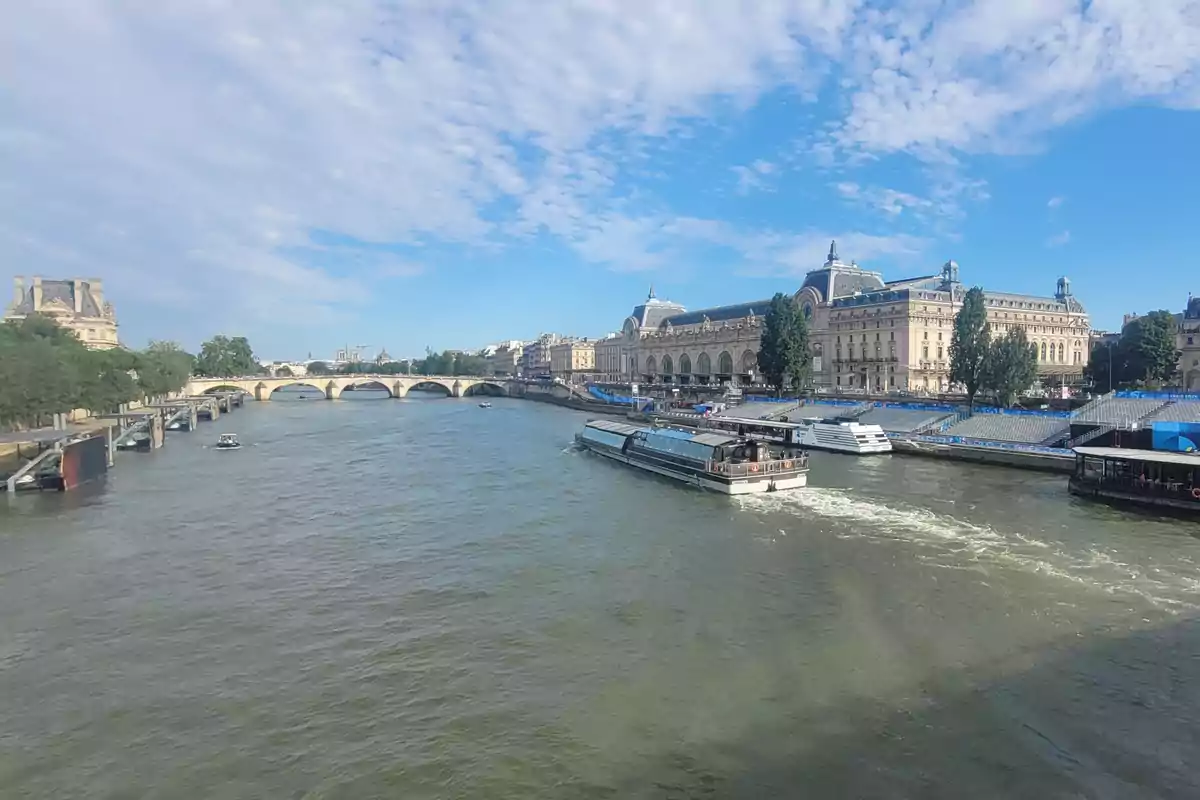 Una vista panorámica de un río con un puente de piedra y edificios históricos a ambos lados, con un barco navegando por el agua bajo un cielo parcialmente nublado.