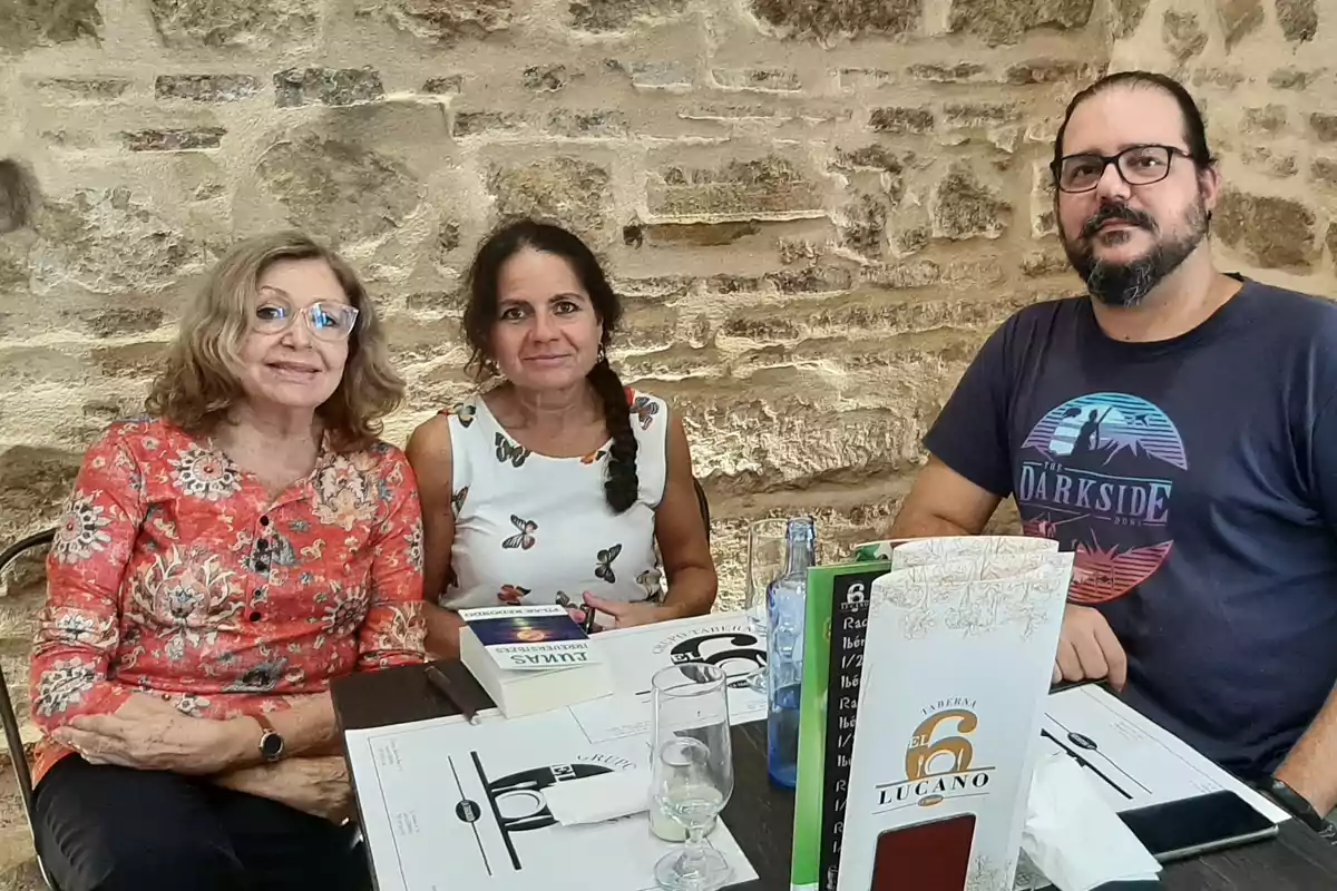 Tres personas están sentadas en una mesa de restaurante con paredes de piedra, hay menús y vasos sobre la mesa.