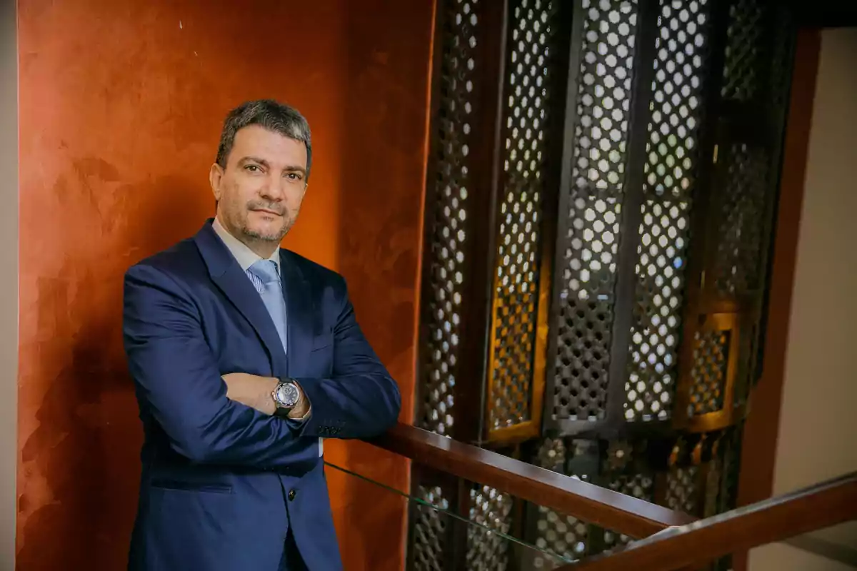 Hombre de traje azul con los brazos cruzados apoyado en una barandilla junto a una pared de color marrón y un fondo de celosía metálica