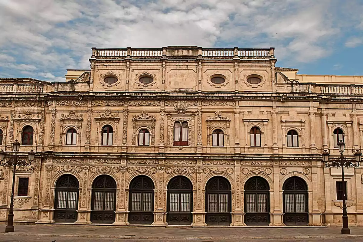 Fachada de un edificio histórico de piedra con detalles ornamentales y ventanas arqueadas bajo un cielo parcialmente nublado