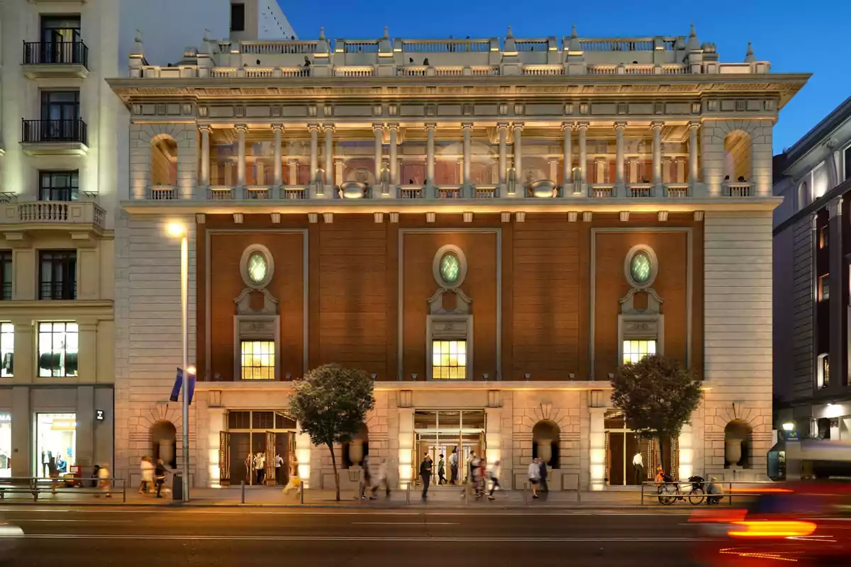 Fachada de un edificio histórico iluminado al anochecer con personas caminando por la acera y árboles frente a la entrada