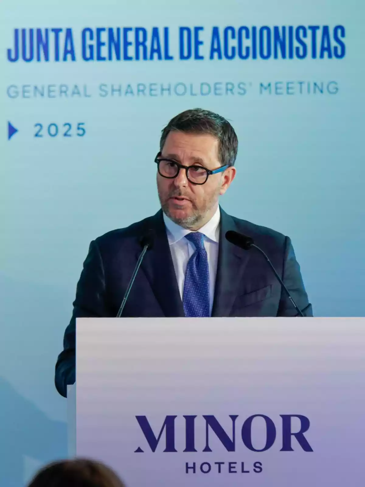 Hombre con gafas y traje hablando en un podio durante la junta general de accionistas de Minor Hotels en 2025 Hombre con gafas y traje hablando en un podio durante la junta general de accionistas de Minor Hotels en 2025