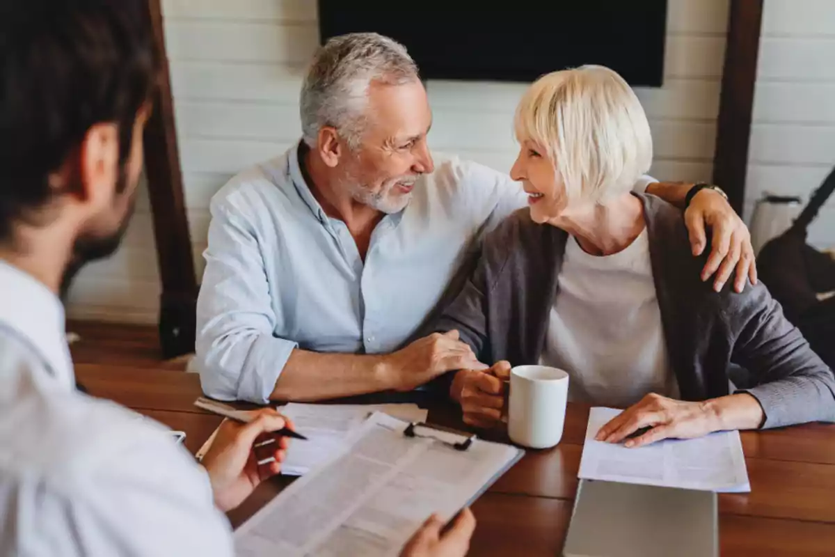 Senior couple smiles while talking with an advisor in an office, surrounded by documents and a white mug.