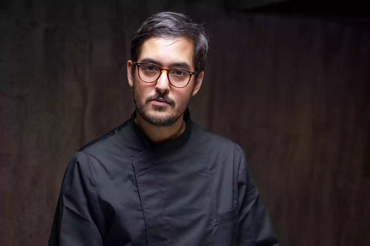 Hombre joven con gafas y barba corta vistiendo un uniforme de chef negro frente a un fondo oscuro Hombre joven con gafas y barba corta vistiendo un uniforme de chef negro frente a un fondo oscuro