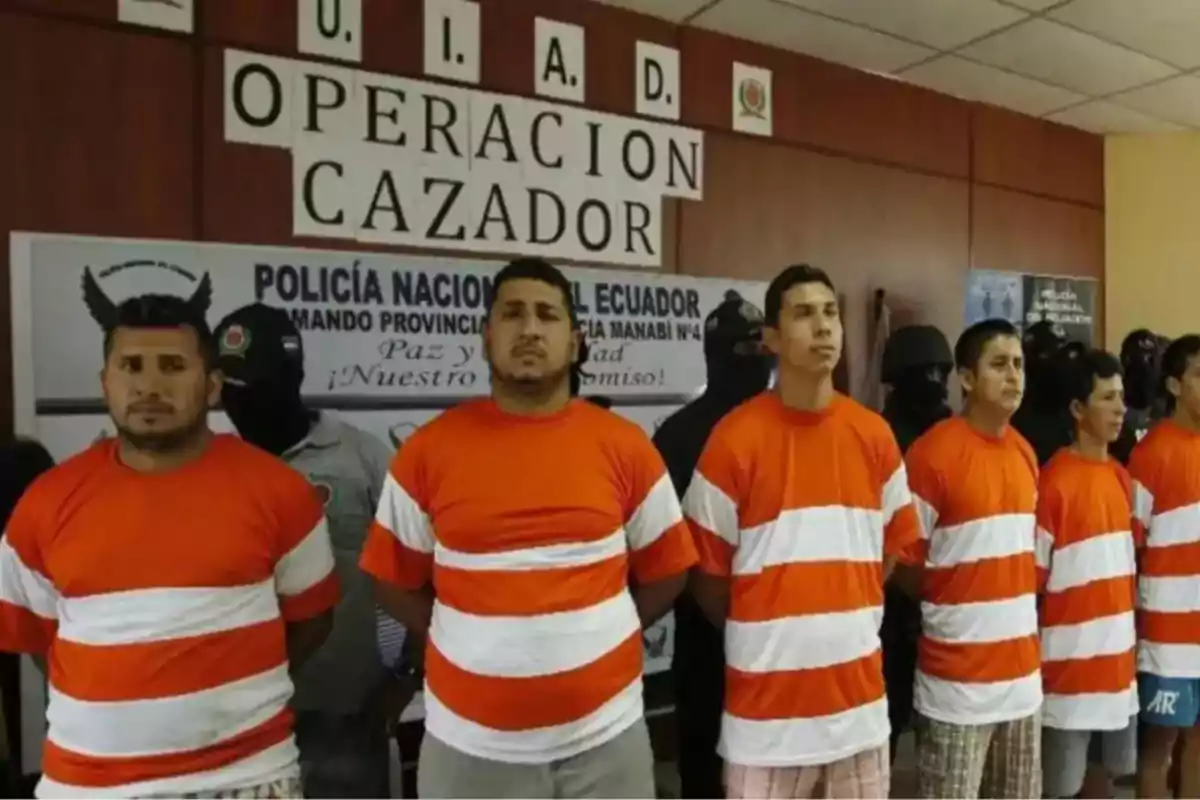 Varios hombres con camisetas naranjas y blancas están alineados frente a un cartel que dice Operación Cazador en una comisaría de policía en Ecuador Varios hombres con camisetas naranjas y blancas están alineados frente a un cartel que dice Operación Cazador en una comisaría de policía en Ecuador