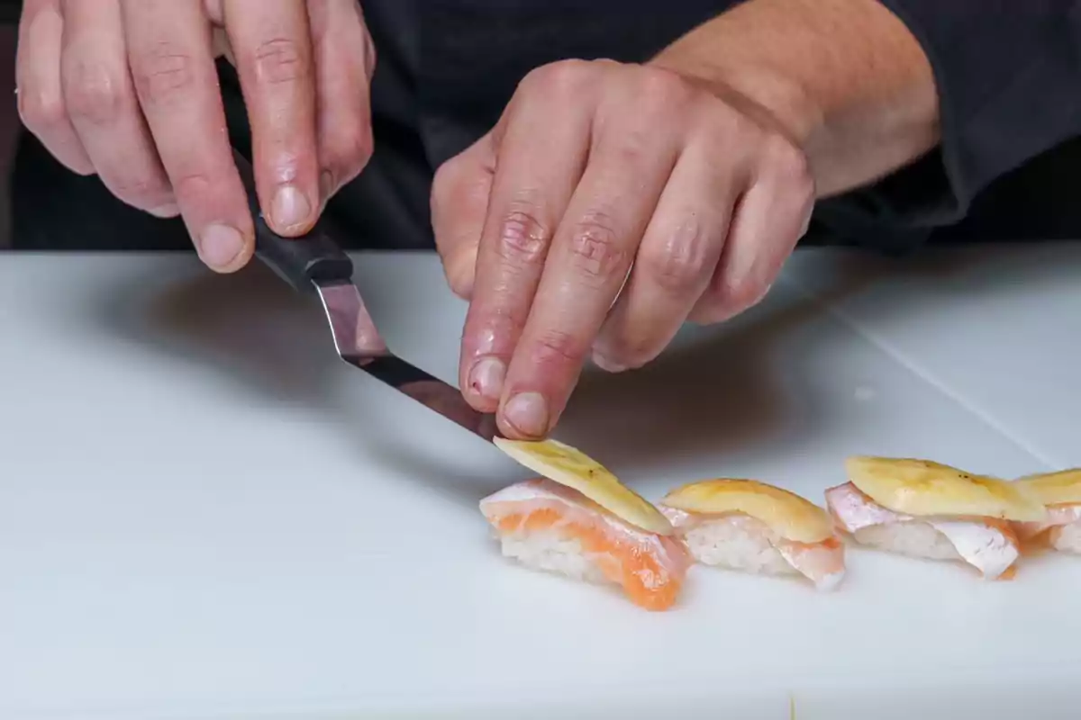 Manos de una persona colocando una lámina de jengibre sobre piezas de sushi en una tabla blanca Manos de una persona colocando una lámina de jengibre sobre piezas de sushi en una tabla blanca
