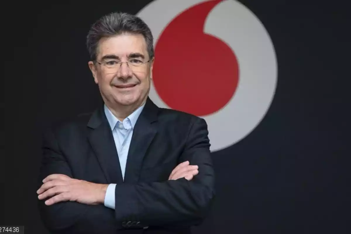 Hombre de cabello canoso y gafas con traje oscuro y camisa azul claro posando con los brazos cruzados frente a un logotipo grande de Vodafone sobre fondo negro Hombre de cabello canoso y gafas con traje oscuro y camisa azul claro posando con los brazos cruzados frente a un logotipo grande de Vodafone sobre fondo negro
