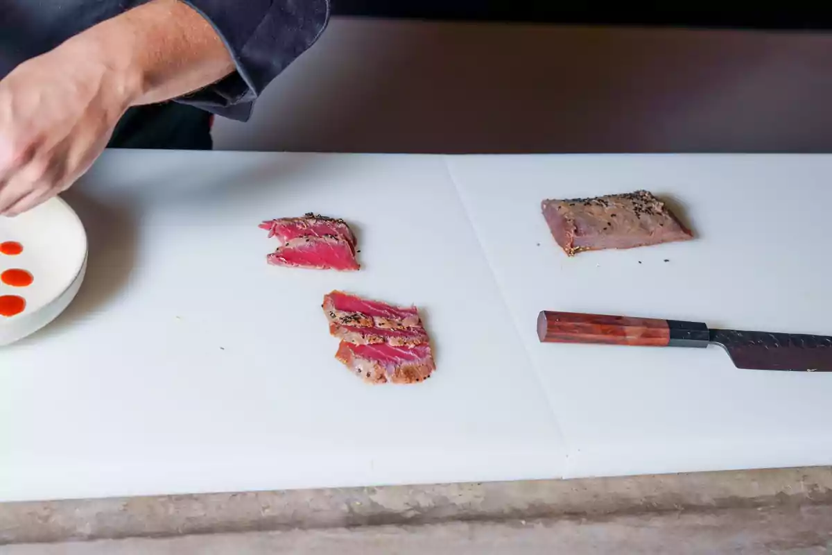 Persona preparando un platillo con atún sellado en una tabla de cortar blanca junto a un cuchillo de mango de madera Persona preparando un platillo con atún sellado en una tabla de cortar blanca junto a un cuchillo de mango de madera