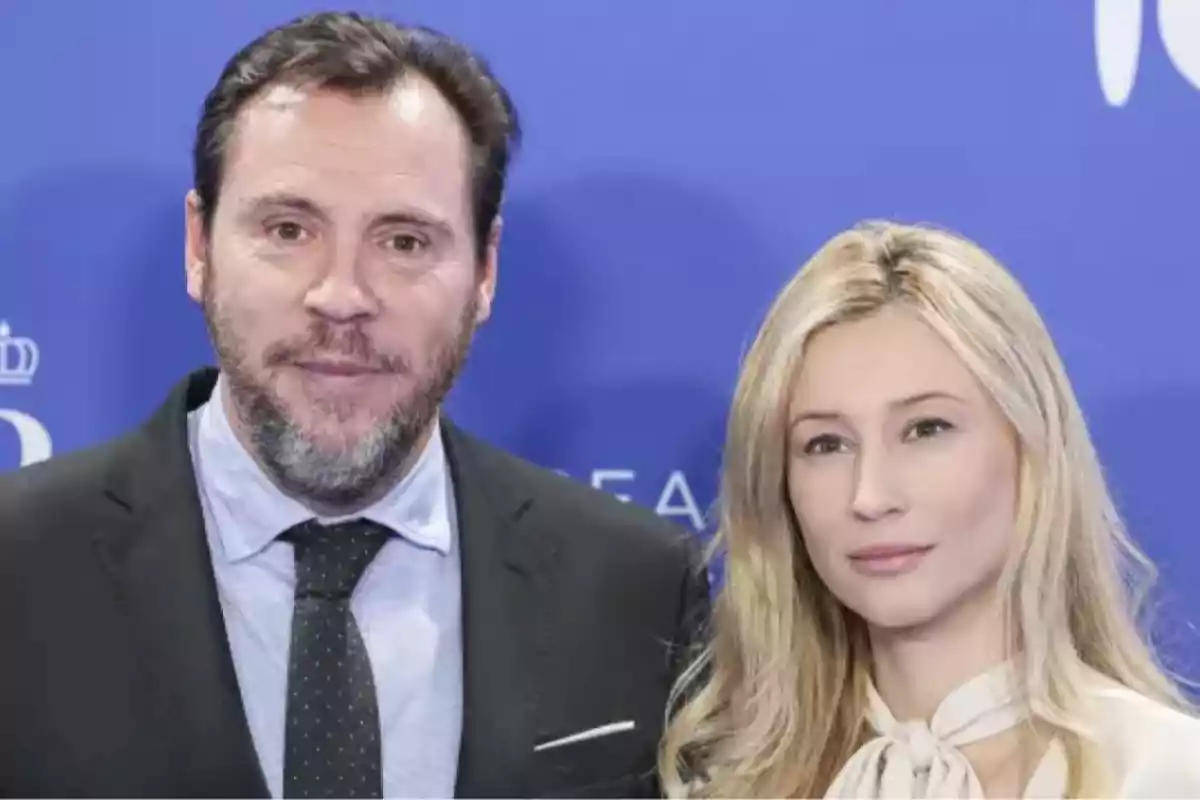 Un hombre con barba y traje oscuro junto a una mujer rubia con blusa clara posan frente a un fondo azul Un hombre con barba y traje oscuro junto a una mujer rubia con blusa clara posan frente a un fondo azul
