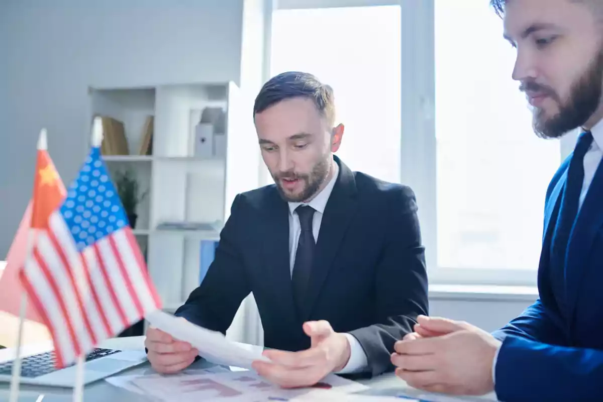 Dos hombres de negocios vestidos con traje revisan documentos en una oficina con banderas de Estados Unidos y China sobre la mesa Dos hombres de negocios vestidos con traje revisan documentos en una oficina con banderas de Estados Unidos y China sobre la mesa