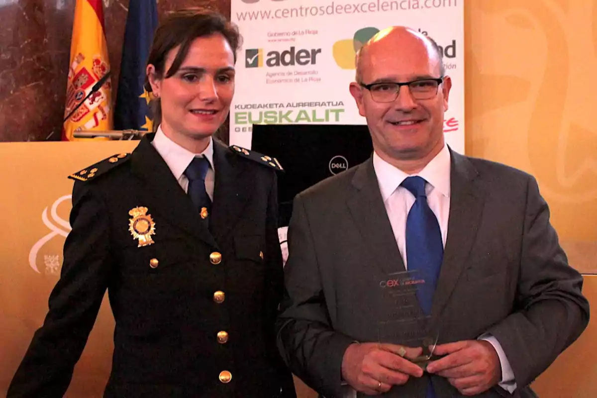 Una mujer con uniforme de policía y un hombre con traje y corbata azul posan juntos en un evento de premiación, el hombre sostiene un trofeo y al fondo se ven banderas y carteles institucionales. Una mujer con uniforme de policía y un hombre con traje y corbata azul posan juntos en un evento de premiación, el hombre sostiene un trofeo y al fondo se ven banderas y carteles institucionales.