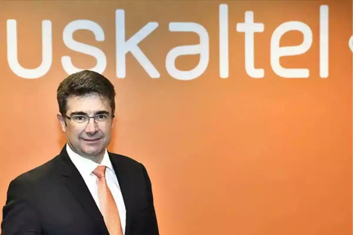 Hombre con gafas y traje oscuro posando frente a una pared naranja con el logo de Euskaltel. Hombre con gafas y traje oscuro posando frente a una pared naranja con el logo de Euskaltel.