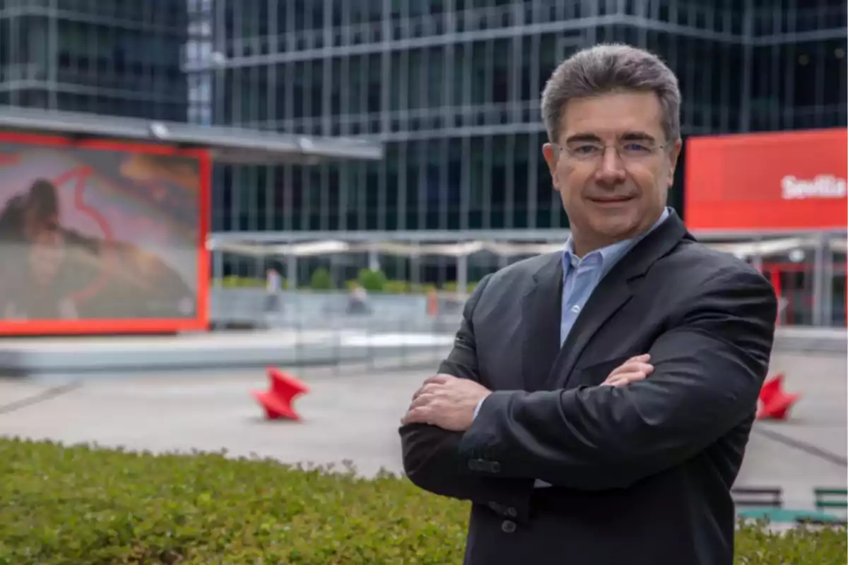 Hombre de cabello canoso y gafas con traje oscuro y camisa azul claro posando con los brazos cruzados en un entorno urbano moderno con edificios de cristal y carteles rojos al fondo Hombre de cabello canoso y gafas con traje oscuro y camisa azul claro posando con los brazos cruzados en un entorno urbano moderno con edificios de cristal y carteles rojos al fondo