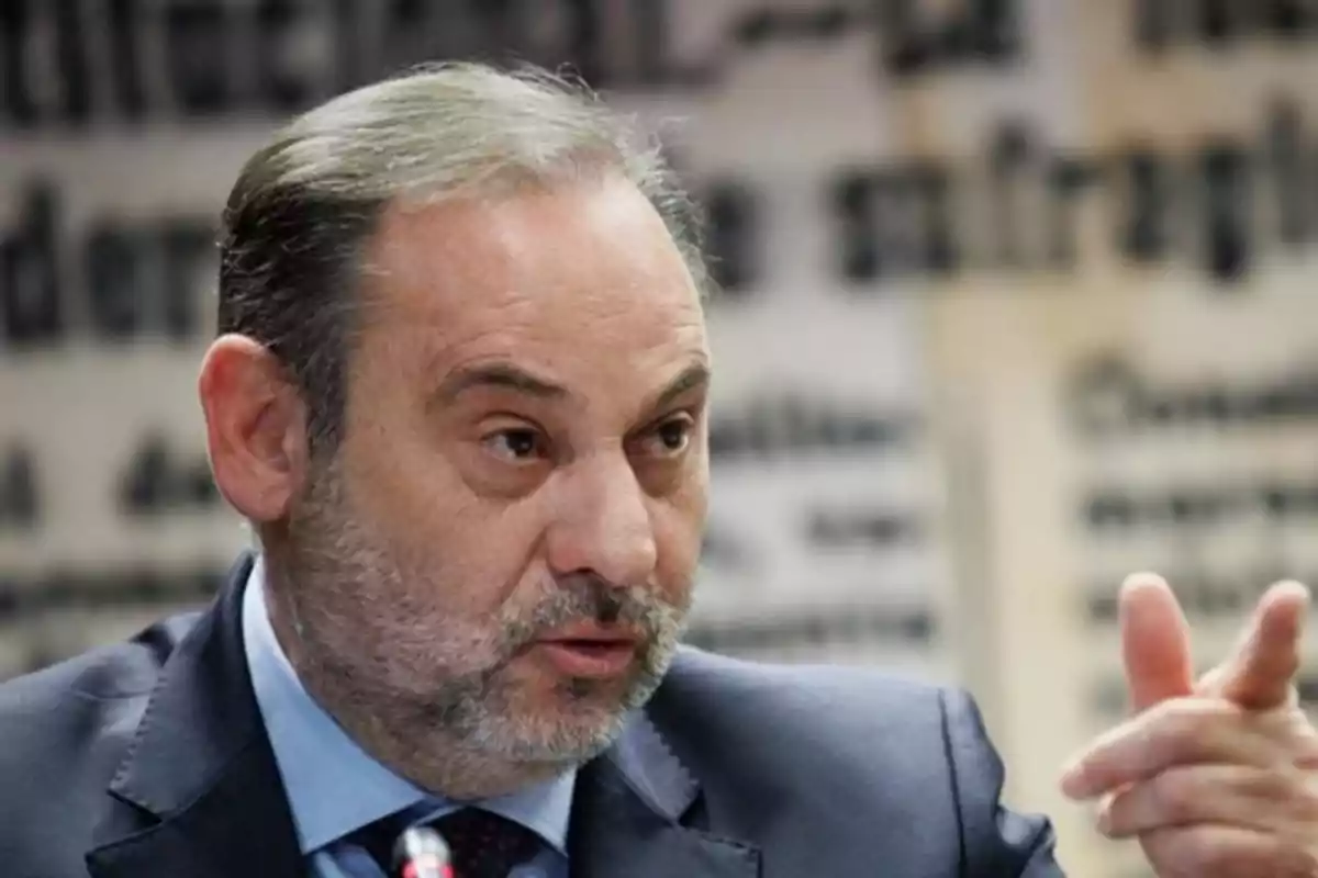 Hombre de mediana edad con barba y cabello corto vestido con traje oscuro hablando y gesticulando con la mano frente a un fondo desenfocado con letras grandes Hombre de mediana edad con barba y cabello corto vestido con traje oscuro hablando y gesticulando con la mano frente a un fondo desenfocado con letras grandes
