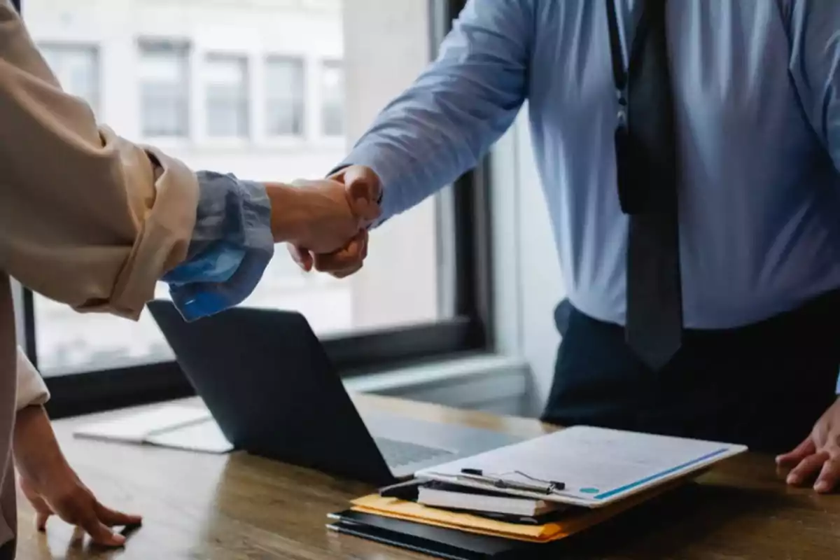 Dos personas dándose la mano en una oficina junto a un escritorio con una laptop y documentos Dos personas dándose la mano en una oficina junto a un escritorio con una laptop y documentos