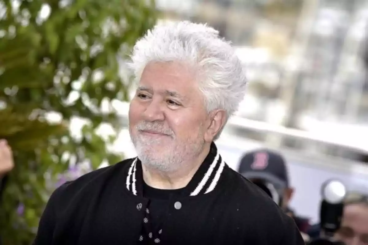 Hombre de cabello canoso y barba corta sonriendo, usando chaqueta negra con detalles blancos, al aire libre con fondo desenfocado de plantas y personas. Hombre de cabello canoso y barba corta sonriendo, usando chaqueta negra con detalles blancos, al aire libre con fondo desenfocado de plantas y personas.