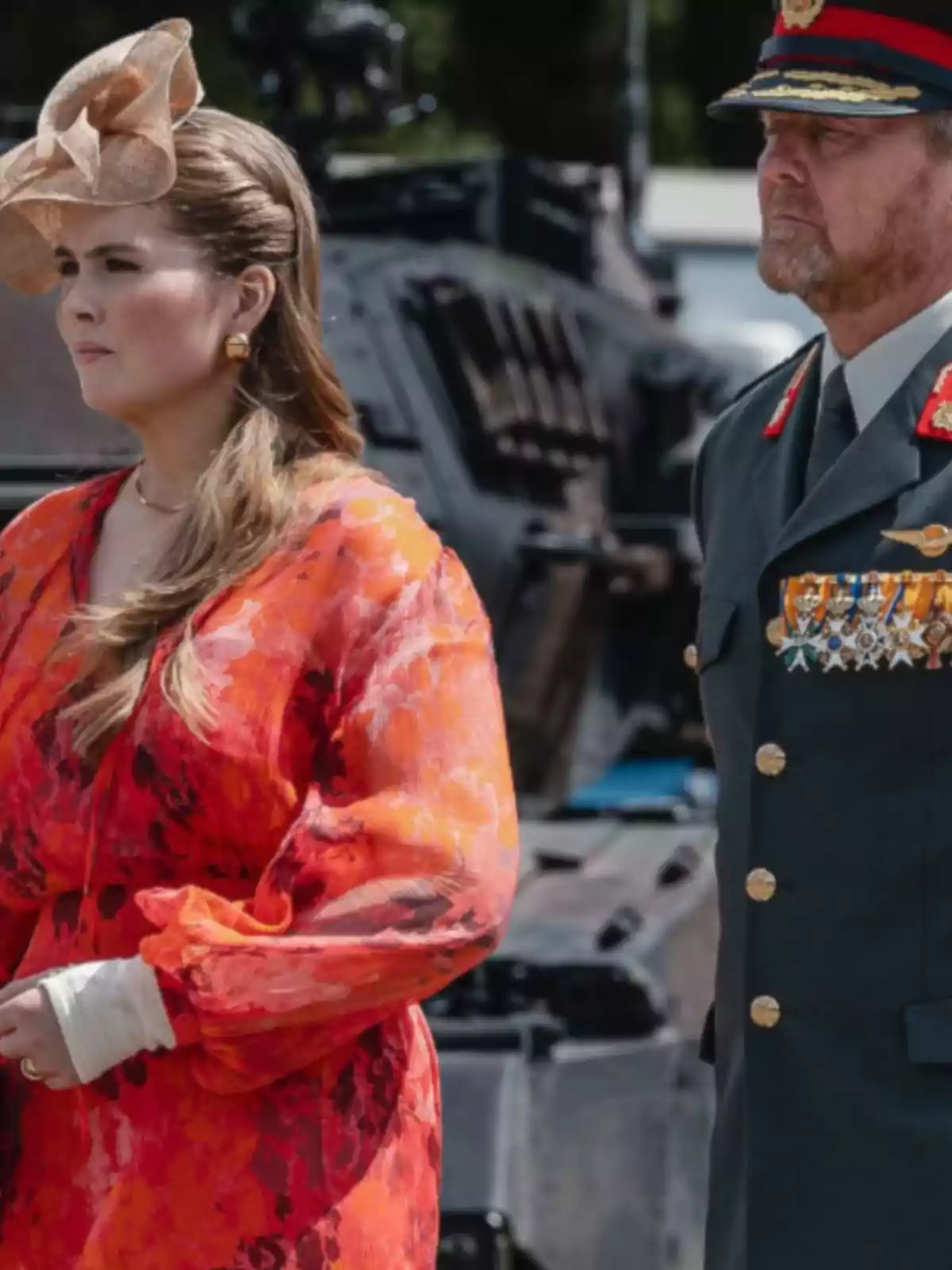 Una mujer con vestido rojo y tocado elegante está junto a un hombre con uniforme militar y medallas, ambos de pie frente a un vehículo blindado. Una mujer con vestido rojo y tocado elegante está junto a un hombre con uniforme militar y medallas, ambos de pie frente a un vehículo blindado.