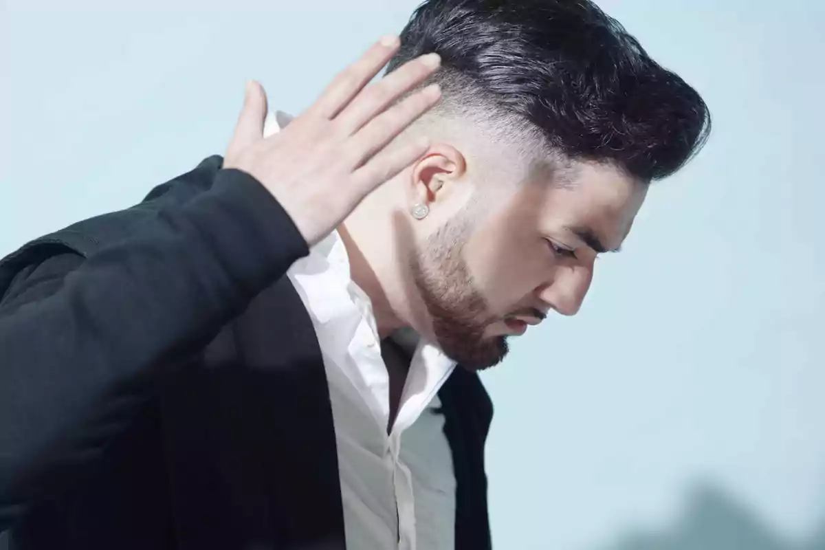 Hombre joven con barba y peinado moderno mirando hacia abajo y levantando una mano