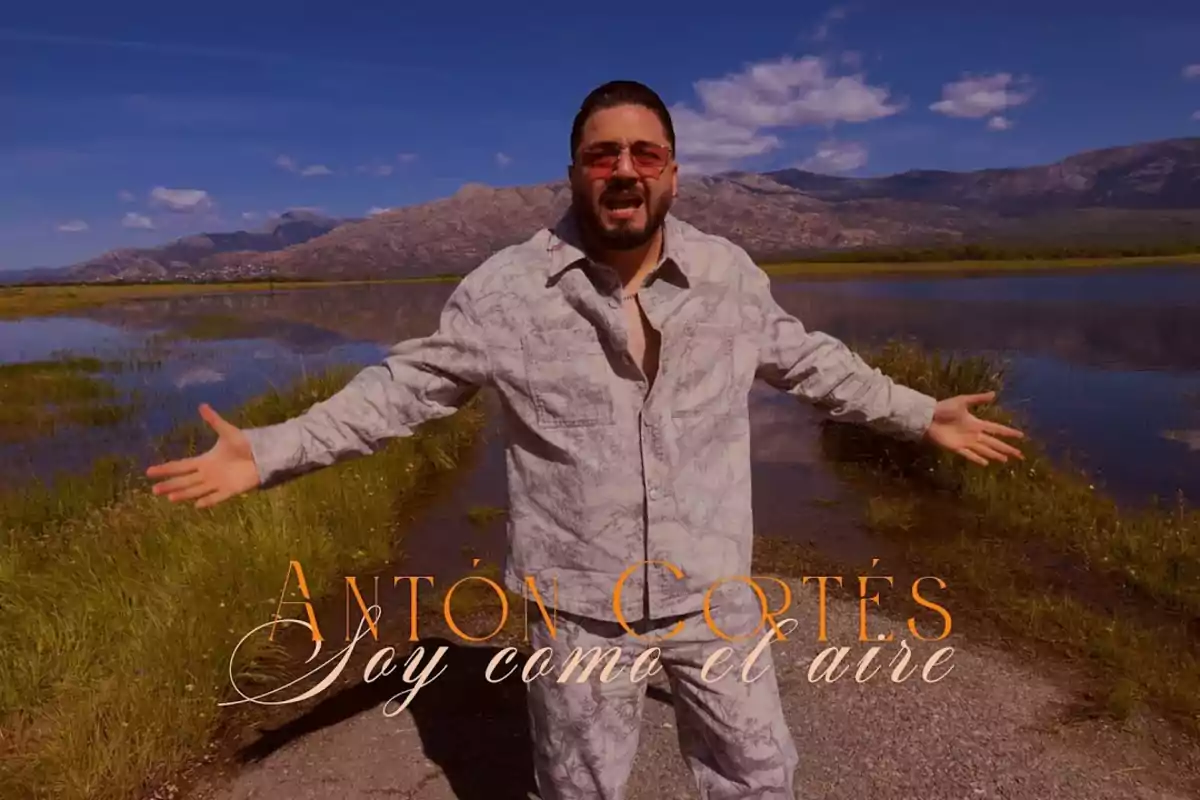 Hombre con gafas de sol y ropa clara posando con los brazos abiertos frente a un paisaje de lago y montañas bajo un cielo azul con el texto Antón Cortés Soy como el aire Hombre con gafas de sol y ropa clara posando con los brazos abiertos frente a un paisaje de lago y montañas bajo un cielo azul con el texto Antón Cortés Soy como el aire