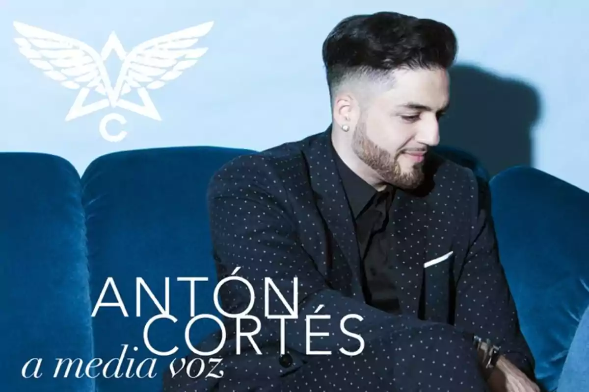 Hombre con barba y cabello oscuro sentado en un sofá azul, vestido con traje negro de puntos, junto al texto Antón Cortés a media voz y un logotipo de alas al fondo. Hombre con barba y cabello oscuro sentado en un sofá azul, vestido con traje negro de puntos, junto al texto Antón Cortés a media voz y un logotipo de alas al fondo.