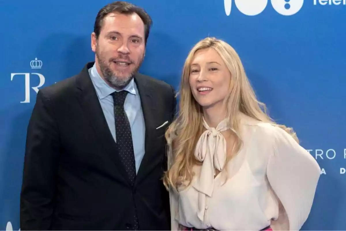 Un hombre con barba y una mujer rubia posan sonrientes frente a un fondo azul en un evento formal. Un hombre con barba y una mujer rubia posan sonrientes frente a un fondo azul en un evento formal.