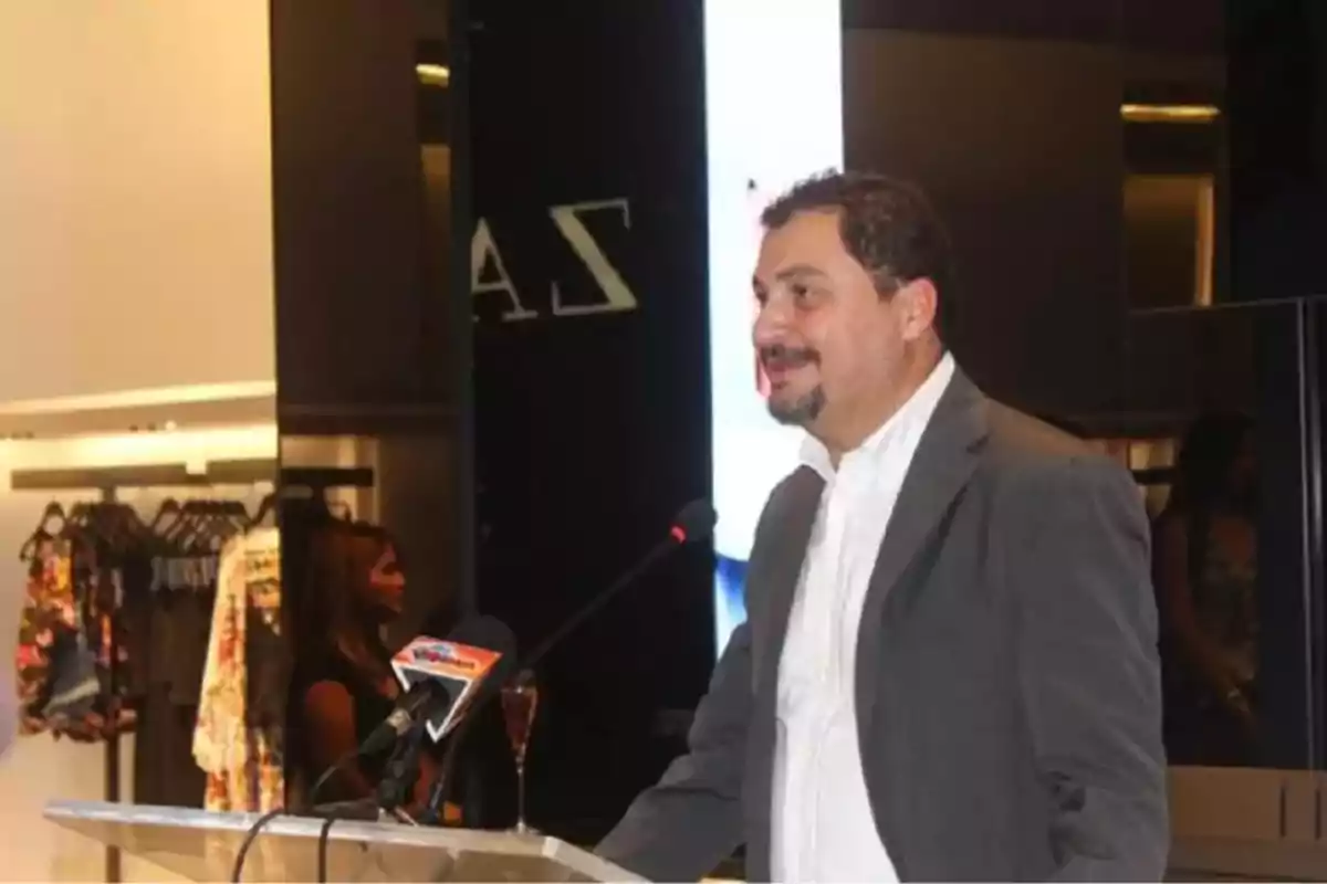 Hombre con barba y saco gris hablando en un micrófono durante un evento en una tienda de ropa