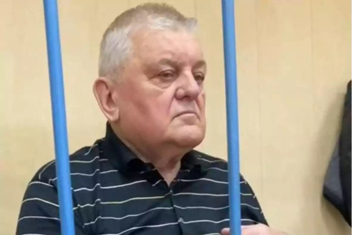 Older man with gray hair and a serious expression sitting behind blue bars, wearing a dark striped jersey in an indoor setting.