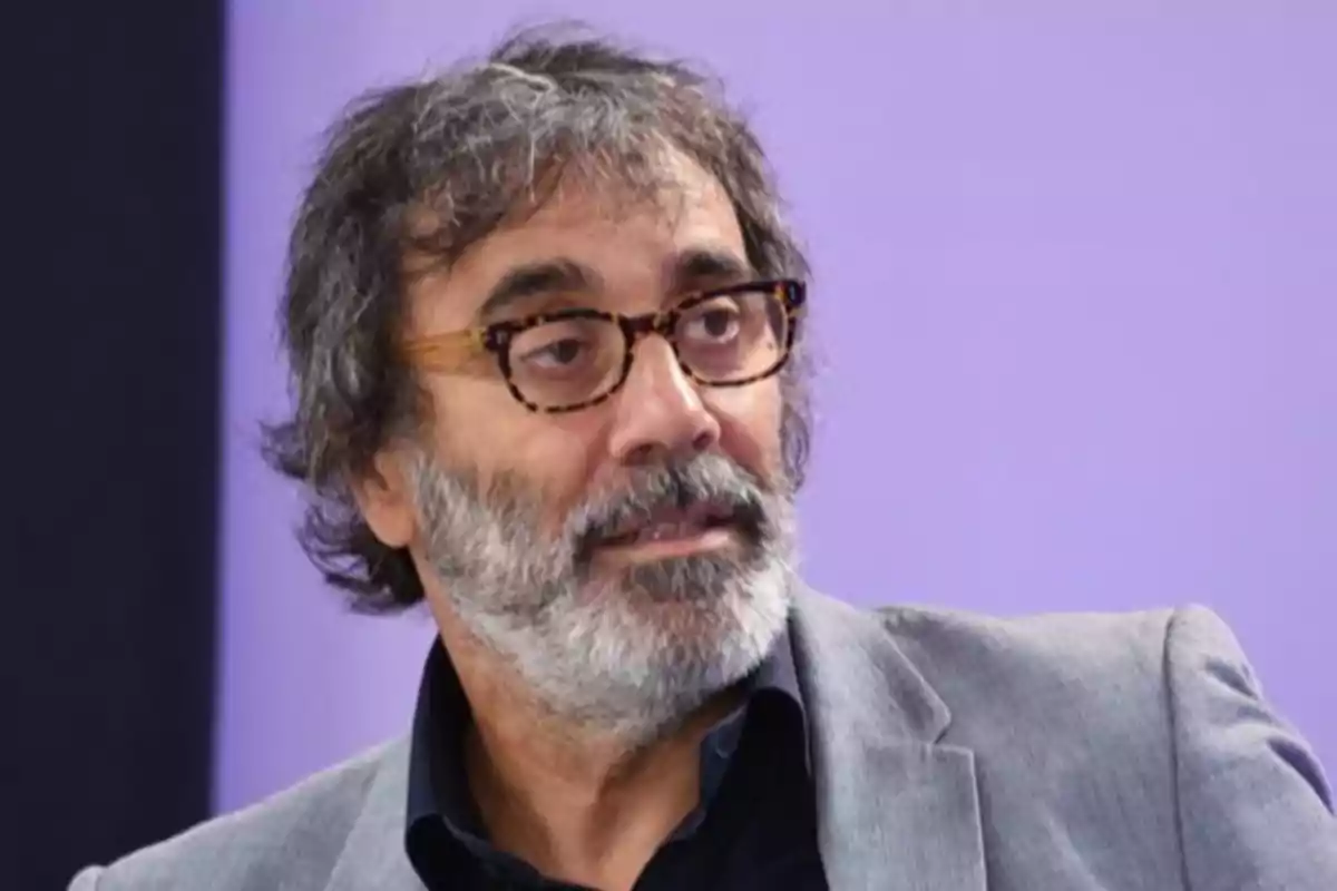 Hombre de cabello canoso y barba con gafas de montura marrón y saco gris frente a un fondo lila
