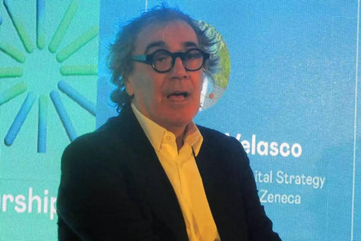 Hombre de cabello canoso y gafas oscuras hablando en un evento con fondo azul y verde con texto y gráficos. Hombre de cabello canoso y gafas oscuras hablando en un evento con fondo azul y verde con texto y gráficos.