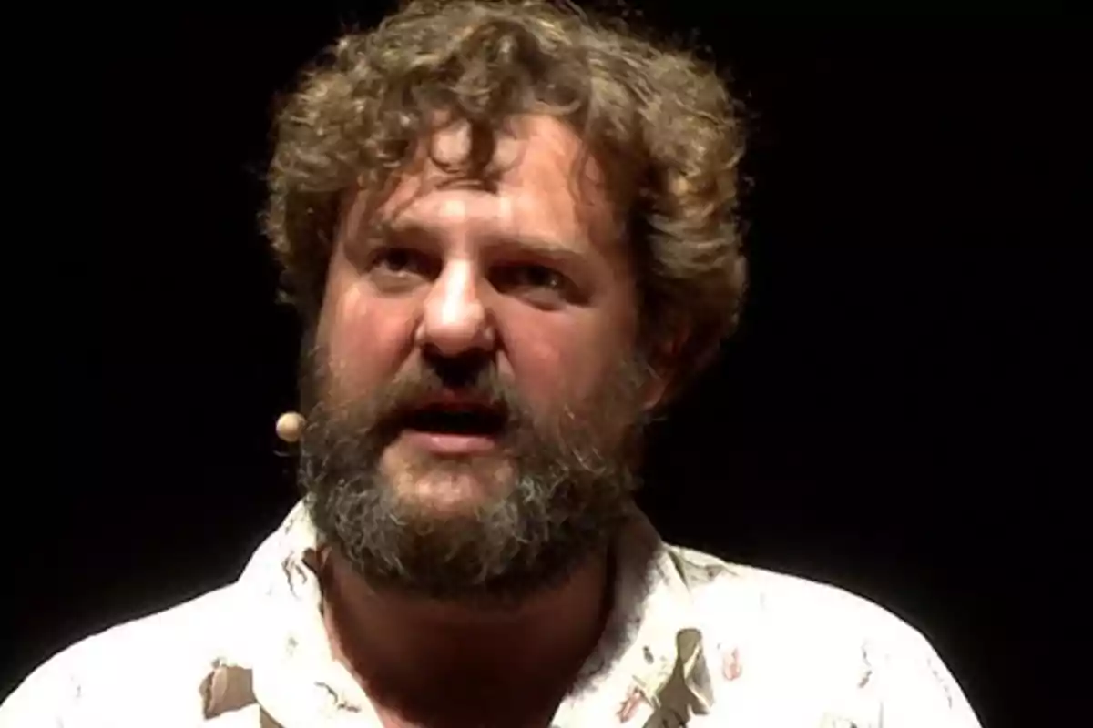 Hombre de cabello rizado y barba con camisa clara hablando con un micrófono de diadema en un fondo oscuro Hombre de cabello rizado y barba con camisa clara hablando con un micrófono de diadema en un fondo oscuro