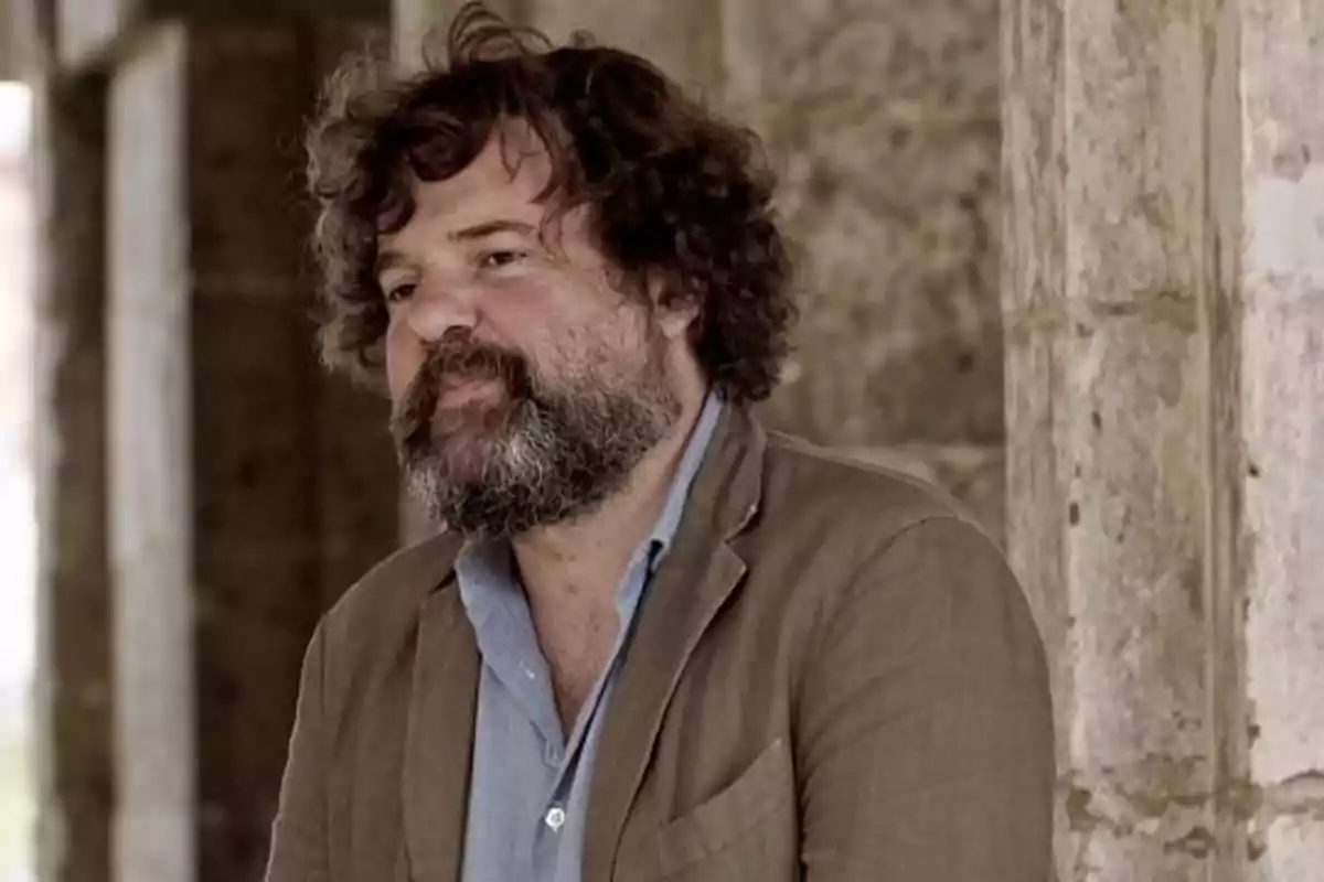 Hombre de cabello rizado y barba con chaqueta marrón y camisa azul claro de pie junto a una pared de piedra