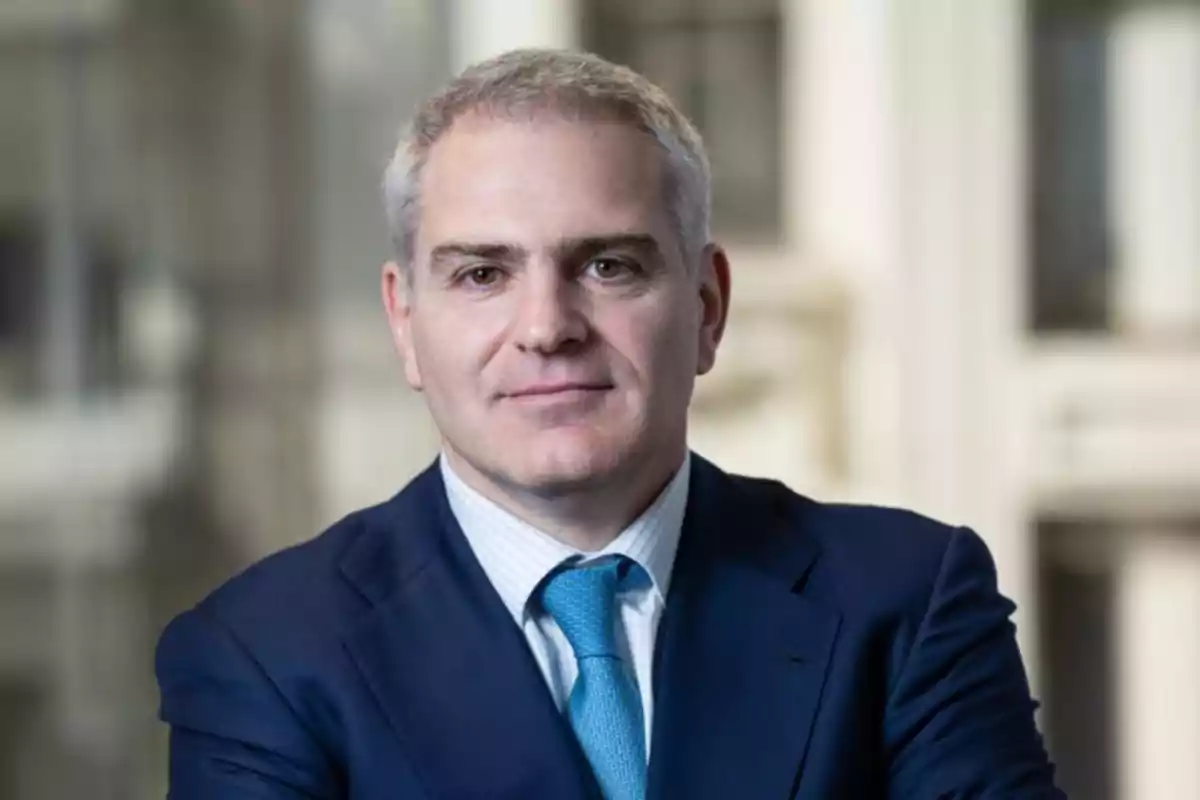 Hombre de cabello canoso con traje azul y corbata celeste posando frente a una ventana en un entorno profesional Hombre de cabello canoso con traje azul y corbata celeste posando frente a una ventana en un entorno profesional
