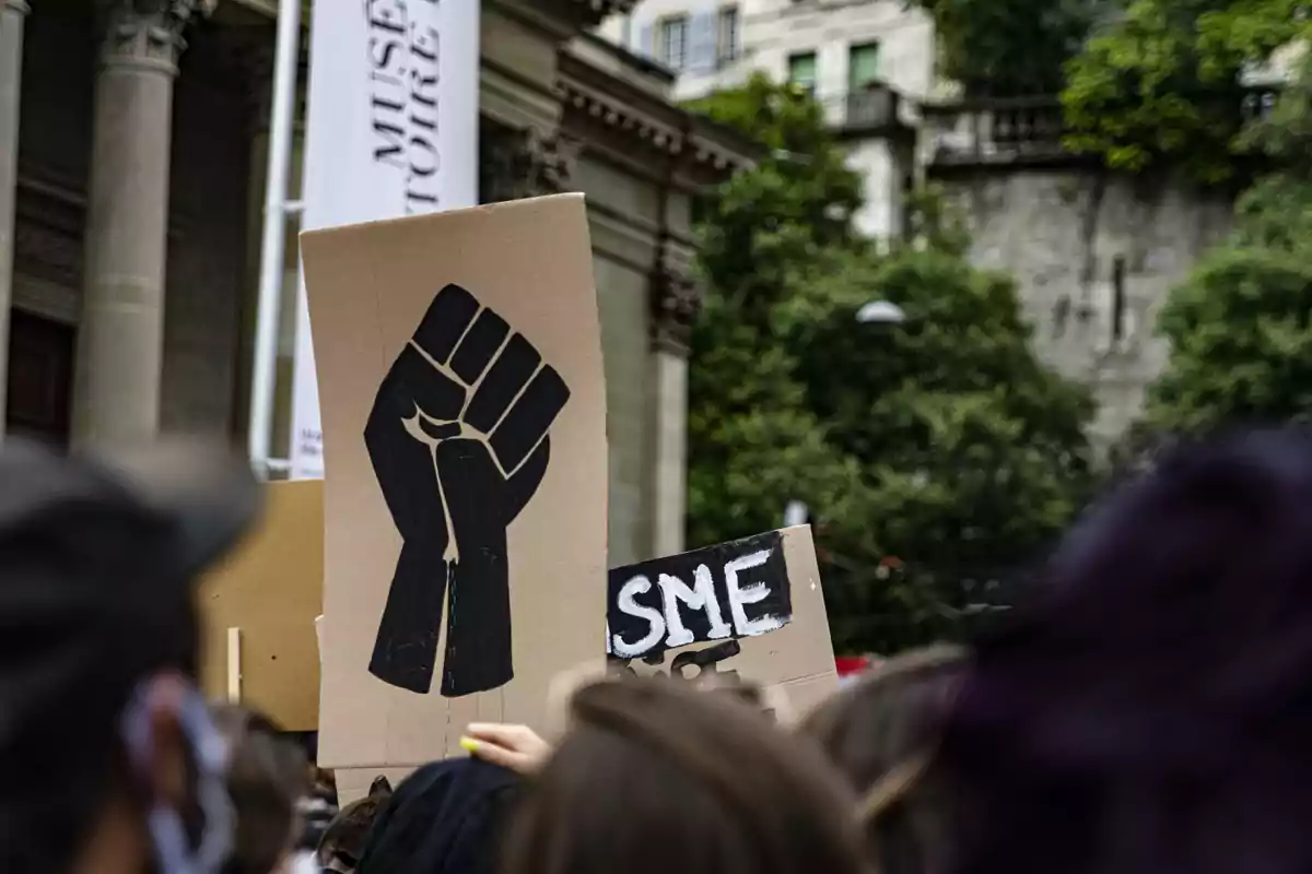 Personas manifestándose con pancartas, una de ellas muestra un puño negro en alto como símbolo de lucha y resistencia Personas manifestándose con pancartas, una de ellas muestra un puño negro en alto como símbolo de lucha y resistencia