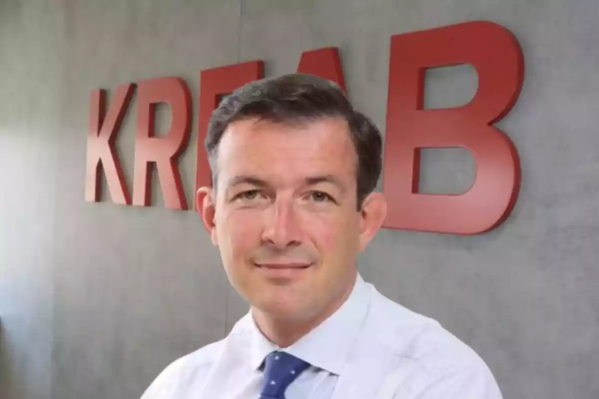 Hombre de cabello corto y camisa clara con corbata azul posando frente a una pared gris con letras rojas grandes que forman la palabra KREAB