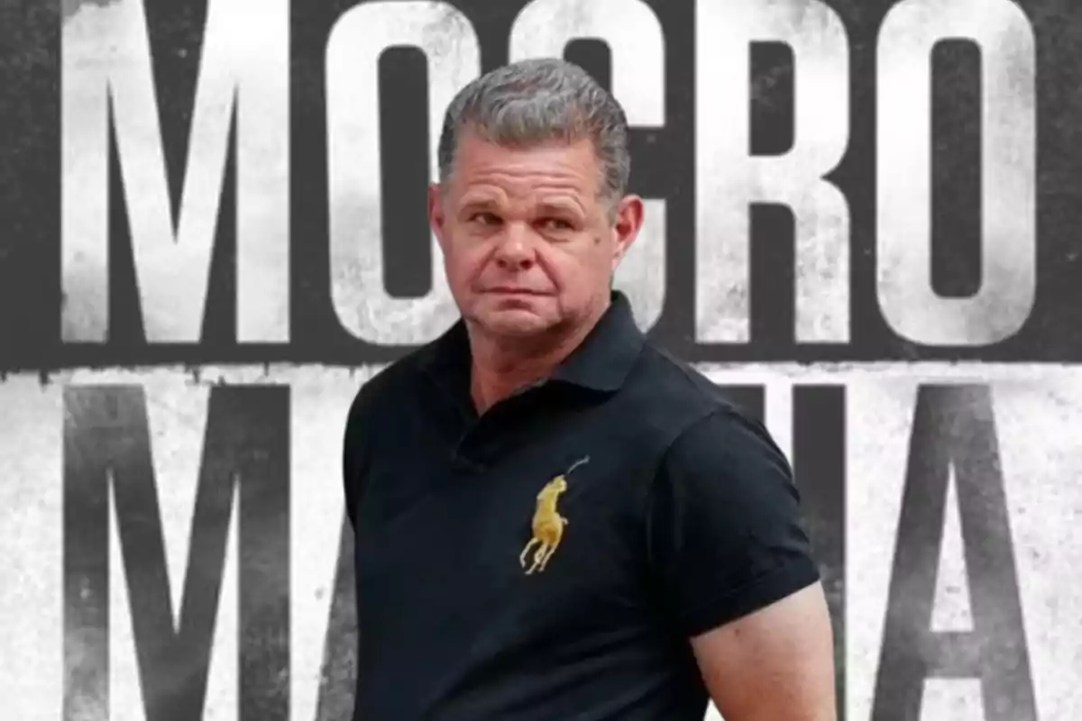 Hombre de cabello corto y canoso con camiseta negra de manga corta y logotipo amarillo, de pie frente a un fondo con letras grandes en blanco y negro.