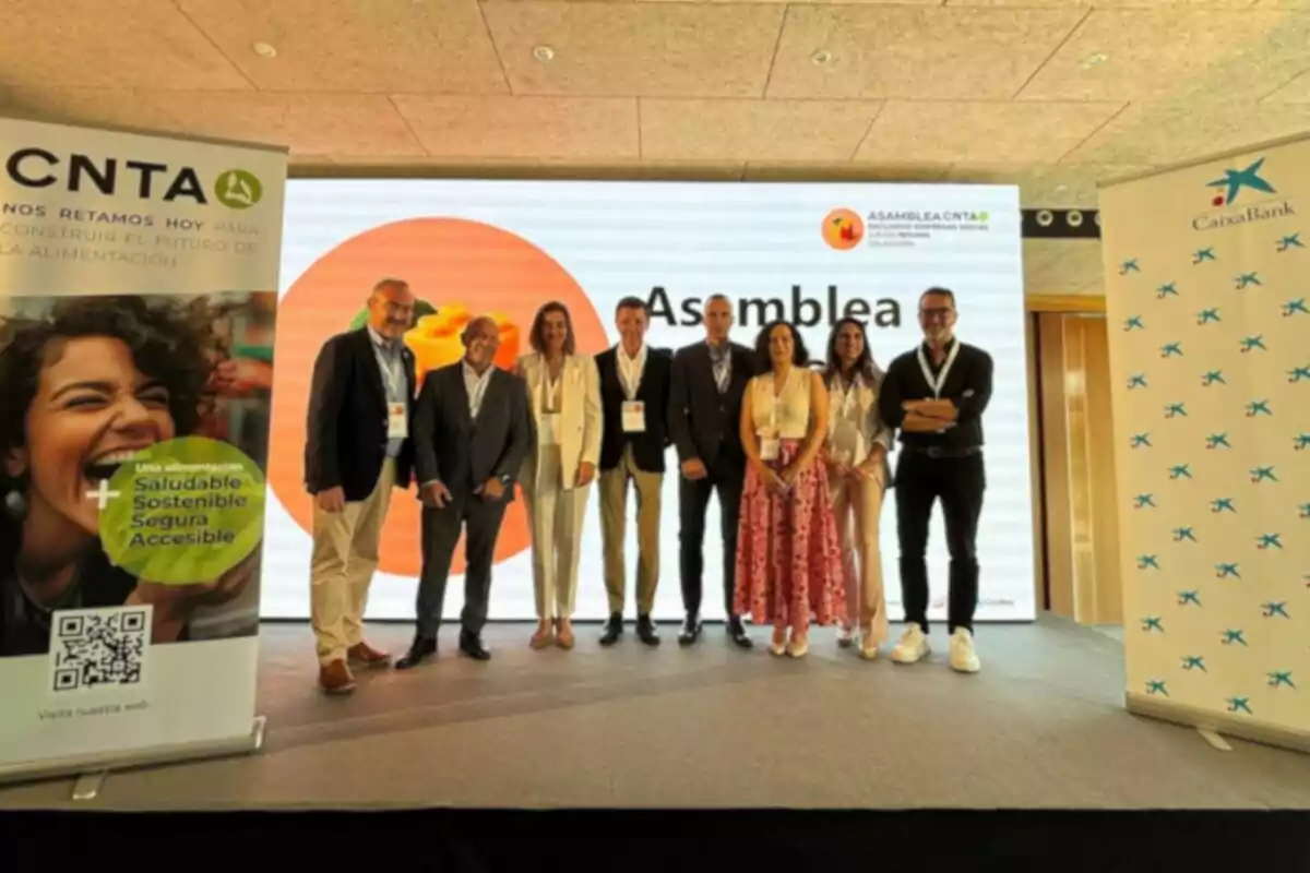 Un grupo de personas posando en un escenario durante un evento corporativo con pancartas de CNTA y CaixaBank a los lados y una pantalla grande de fondo que muestra la palabra Asamblea Un grupo de personas posando en un escenario durante un evento corporativo con pancartas de CNTA y CaixaBank a los lados y una pantalla grande de fondo que muestra la palabra Asamblea