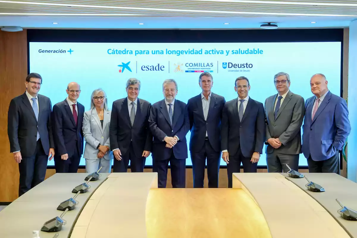 Grupo de personas vestidas de traje posando frente a una pantalla que muestra el texto Cátedra para una longevidad activa y saludable junto a los logotipos de varias instituciones educativas y una mesa de reuniones en primer plano