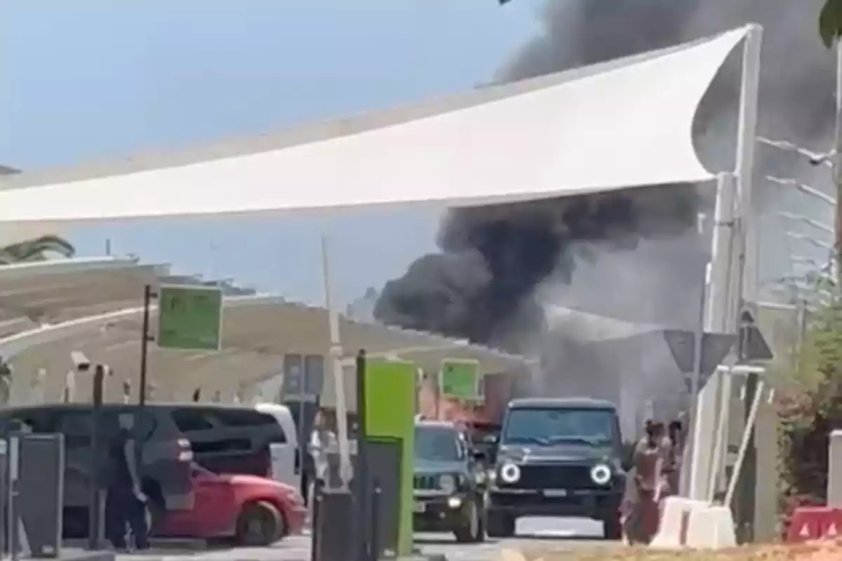 Varios autos estacionados bajo una estructura blanca mientras una columna de humo negro se eleva al fondo Varios autos estacionados bajo una estructura blanca mientras una columna de humo negro se eleva al fondo