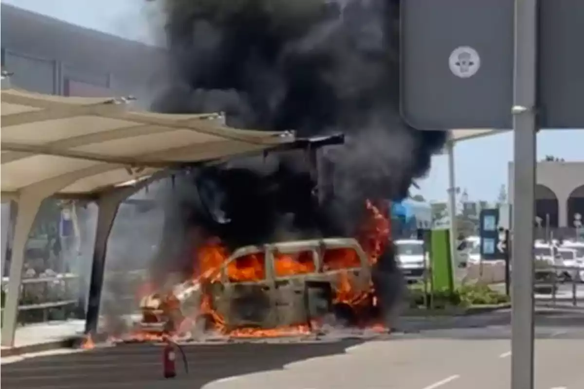 Una camioneta envuelta en llamas con humo negro saliendo de ella en una zona urbana junto a una estructura de sombra y un extintor en el suelo. Una camioneta envuelta en llamas con humo negro saliendo de ella en una zona urbana junto a una estructura de sombra y un extintor en el suelo.