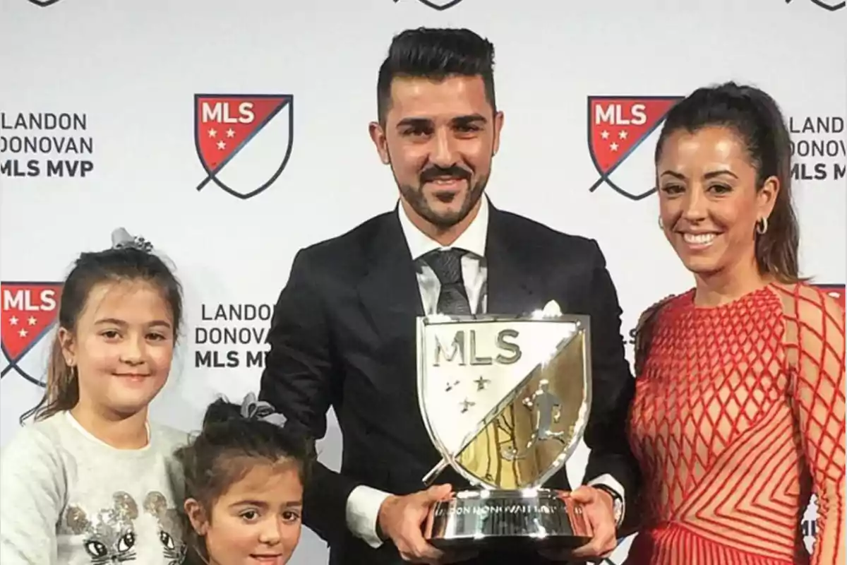 Un hombre con traje sostiene un trofeo de la MLS y posa sonriente junto a dos niñas y una mujer frente a un fondo con logotipos de la liga Un hombre con traje sostiene un trofeo de la MLS y posa sonriente junto a dos niñas y una mujer frente a un fondo con logotipos de la liga