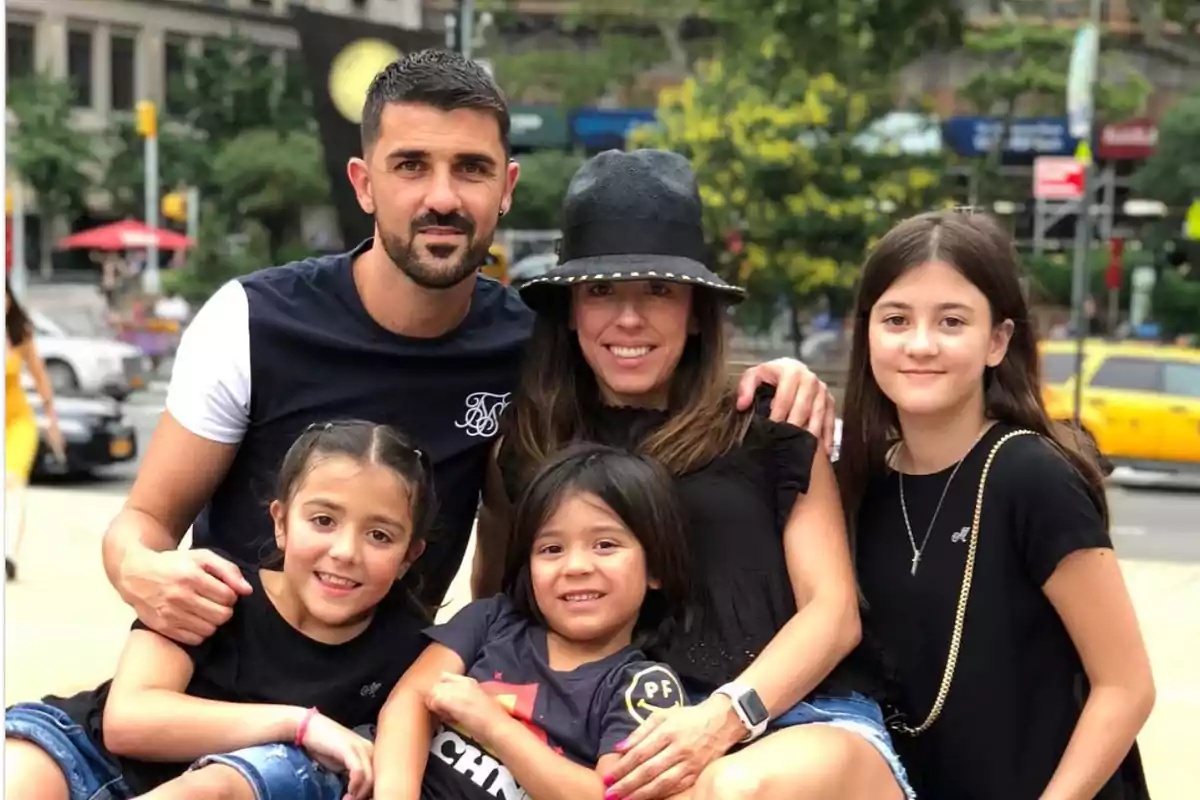 Una familia de cinco personas sonríe mientras posa sentada al aire libre en una ciudad con árboles y autos al fondo Una familia de cinco personas sonríe mientras posa sentada al aire libre en una ciudad con árboles y autos al fondo
