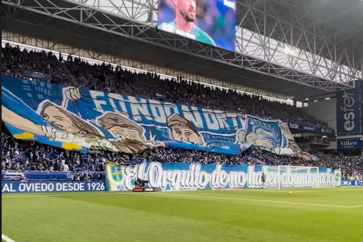 Aficionados en un estadio de fútbol despliegan una gran pancarta azul con dibujos y mensajes de apoyo al equipo, mientras la grada está llena de seguidores animando. Aficionados en un estadio de fútbol despliegan una gran pancarta azul con dibujos y mensajes de apoyo al equipo, mientras la grada está llena de seguidores animando.